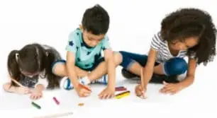 Tres niños dibujando y trabajando con colores sobre una mesa blanca.