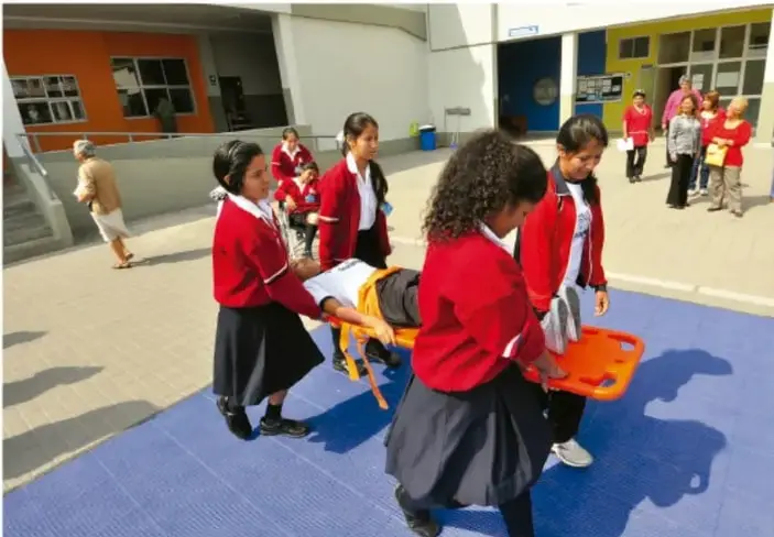 Fotografía de estudiantes de primaria con uniforme realizando un simulacro: trasladan a una compañera en una camilla rígida naranja sobre una superficie azul, en el patio de la escuela.