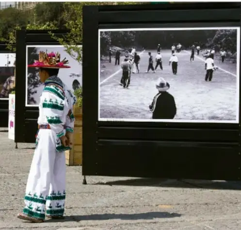 Mujer indígena con vestimenta tradicional observando una fotografía en gran formato exhibida al aire libre en el espacio público.