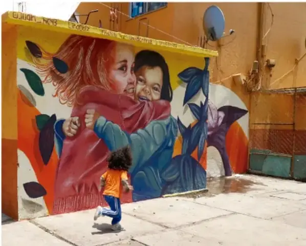 Mural colorido en una esquina de calle que muestra a dos niñas abrazándose con frondas vegetales de fondo; una niña real corre frente al mural. Simboliza la convivencia armónica y el respeto entre pares en el espacio público.