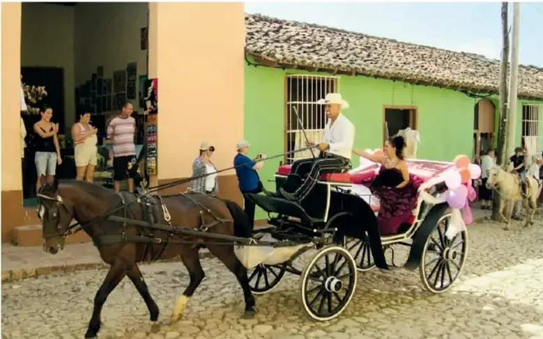 Fotografía de un carruaje tirado por caballo circulando por una calle empedrada de un pueblo; dentro viaja una quinceañera con vestido y globos, acompañada de un cochero con sombrero, mientras vecinos observan desde las banquetas.