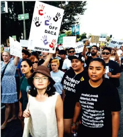 Marcha de personas migrantes con carteles que dicen OFF DACA y mensajes de derechos humanos.