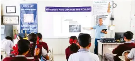 Fotografía de un aula de Telesecundaria con estudiantes frente a una pantalla donde un docente explica contenido educativo; lema visible: 'What starts here, CHANGES THE WORLD'.