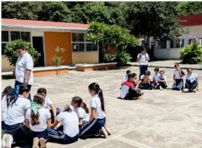 Fotografía de estudiantes de telesecundaria reunidos en círculos en el patio de su escuela, participando en una actividad grupal al aire libre con su maestro.