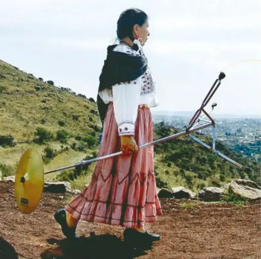 Fotografía de una mujer con vestimenta tradicional (falda rosa larga, blusa bordada, rebozo negro) que carga en la espalda una pieza metálica de arado con disco amarillo, caminando cuesta arriba en un cerro desde el que se ve una ciudad.