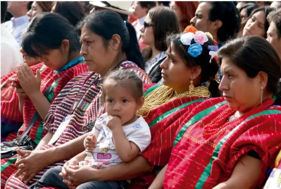 Fotografía de una asamblea comunitaria: grupo de mujeres indígenas con trajes tradicionales coloridos (huipiles rojos) sentadas en filas, una de ellas con una niña pequeña en brazos, participando en una reunión comunitaria.