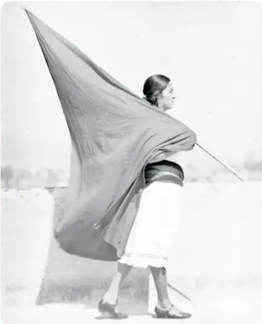 Fotografía en blanco y negro de una mujer de perfil que camina sobre un muro o camino cargando una gran bandera de tela clara que ondea; falda clara y blusa oscura; pie: 'Mujer con bandera, ca. 1928'.