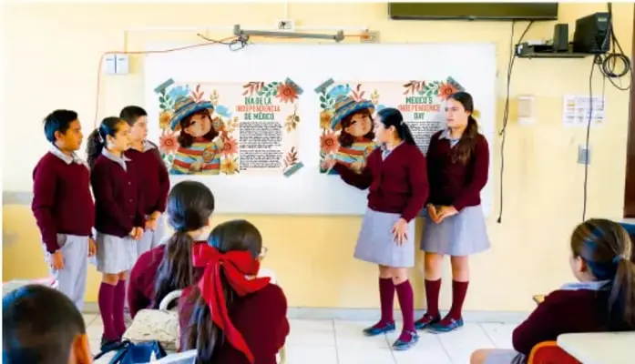 Fotografía de estudiantes de telesecundaria con uniforme realizando una presentación oral frente a un pizarrón con dos carteles ilustrados, mientras sus compañeros observan sentados.