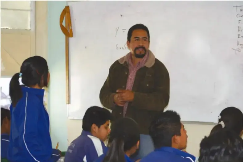 Un docente frente a un grupo de estudiantes en el aula, junto a un pizarrón.