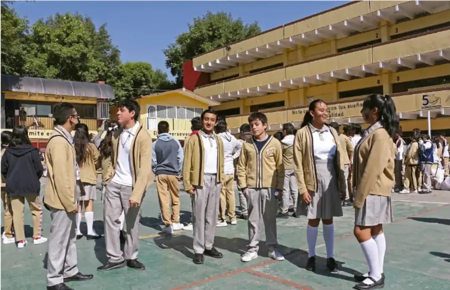 Fotografía de estudiantes de secundaria con uniforme conversando y conviviendo en el patio o cancha de su escuela, con edificios escolares al fondo.