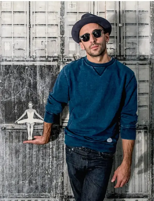 Fotografía del artista JR posando con sombrero y lentes oscuros, sosteniendo en su mano una pequeña figura recortada de una bailarina sobre una pared enrejada.