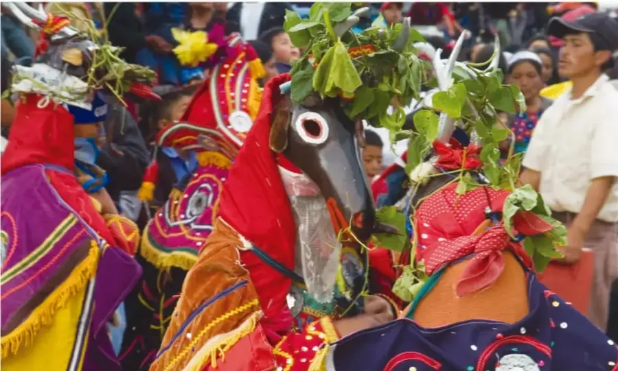 Danzantes tradicionales con trajes multicolores (rojo, morado, amarillo) decorados con hojas naturales y flores, uno con máscara de animal de ojos grandes; alrededor hay espectadores en una fiesta popular.