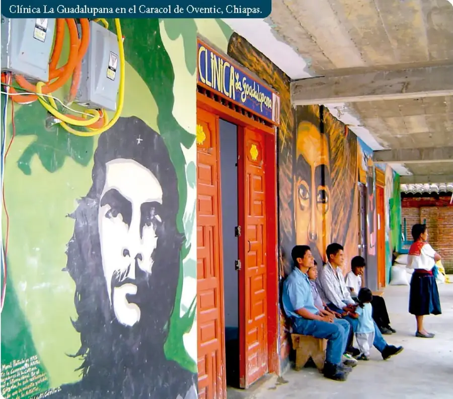 Fotografía de la Clínica La Guadalupana en el Caracol de Oventic, Chiapas. Se observa un edificio con murales, puertas rojas y personas sentadas esperando atención médica.