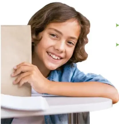 Niña sonriente apoyada en una mesa con un libro, mirando hacia el frente.