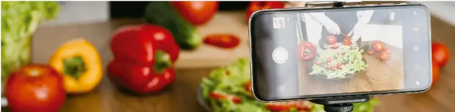 Fotografía de un teléfono celular montado en trípode grabando un video de preparación de una ensalada fresca con verduras como jitomate, pimientos y lechuga sobre una tabla de cocina.