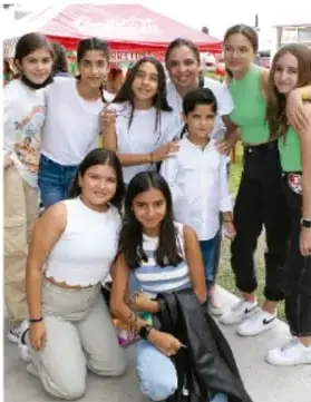 Fotografía de un grupo de adolescentes y niñas posando juntas al aire libre, algunas con camisetas blancas y una con camiseta verde, junto a una mesa con mantel rojo; ilustra la colaboración y el trabajo colectivo.
