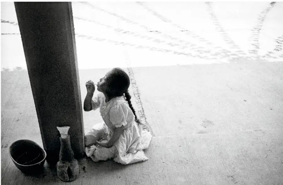 Fotografía en blanco y negro de una niña pequeña con vestido blanco sentada en el piso junto a un poste de madera, con un jarro y un recipiente al lado; en el suelo hay marcas dibujadas.