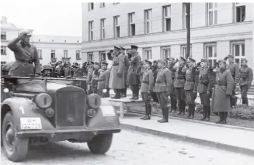 Fotografía en blanco y negro de un desfile militar alemán de la época nazi: soldados formados en fila junto a un automóvil descapotable del que un militar saluda con el brazo extendido.