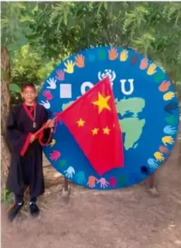 Estudiante de pie junto a una rueda o disco decorado con el mapa del mundo, banderas (incluye la bandera de China) y la palabra 'ODS', en un entorno al aire libre con árboles.