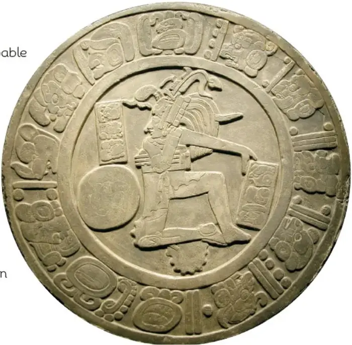 Disco de piedra tallado en relieve con una figura de jugador de pelota sentado o arrodillado junto a una pelota, enmarcado por una orilla decorada con glifos tallados.