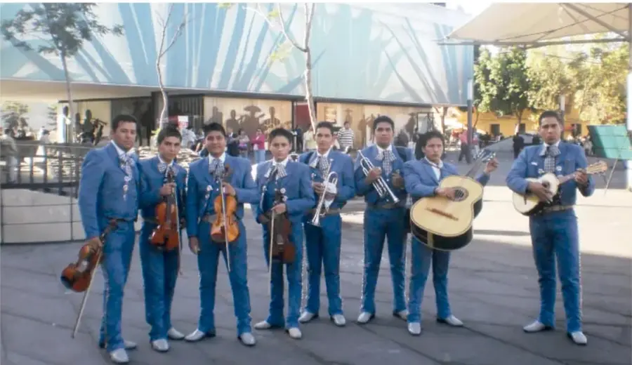Fotografía de un grupo de mariachi con instrumentos de cuerda y viento frente a un edificio moderno con árboles y personas al fondo.
