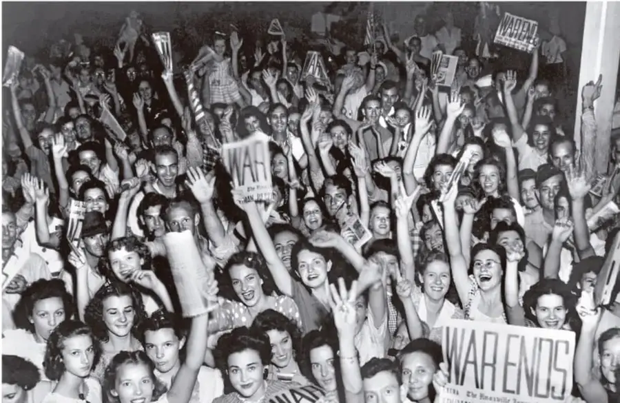 Fotografía en blanco y negro de una multitud celebrando el fin de la Segunda Guerra Mundial. Varias personas sostienen carteles con la palabra 'WAR ENDS' (la guerra termina).