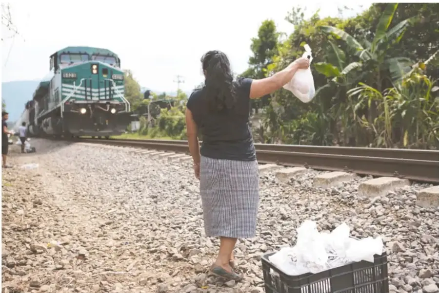 Fotografía de una mujer de espaldas, con falda a cuadros, levantando una bolsa con alimentos frente al tren de carga que avanza por las vías; junto a ella, un cajón plástico negro con bolsas blancas preparadas para entregar.