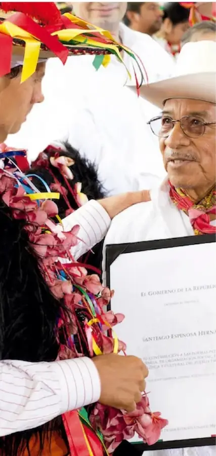 Fotografía de dos adultos mayores en un evento: uno con sombrero y lentes recibe un documento con membrete del Gobierno de la República, rodeados de adornos de flores de colores (confeti) y personas de blanco.