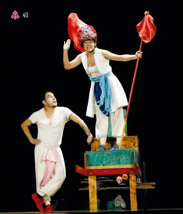 Fotografía de dos actores en escena con vestuario de colores claros y rojo; uno sostiene una bandera roja sobre una tarima de madera pintada de verde y rojo.