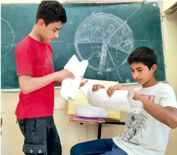 Fotografía de dos estudiantes de telesecundaria frente a un pizarrón con un esquema circular dibujado a tiza, revisando hojas y materiales del proyecto. Crédito: Jesús Eduardo Arteaga Flores, Tamaulipas.