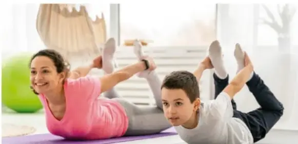 Niña y niño haciendo ejercicio de estiramiento sobre colchonetas, con una pelota verde al fondo.