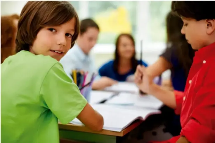 Fotografía de estudiantes de primaria trabajando en equipo en un salón de clases; un niño con camisa verde mira a la cámara y otro con camisa roja escribe.