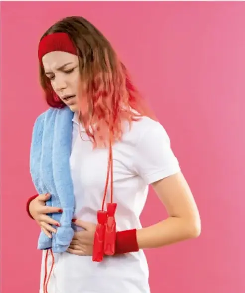 Fotografía de una joven con expression de dolor sosteniéndose el abdomen, con toalla y botella, sobre fondo rosa; ilustra los molestias físicas de la menstruación.