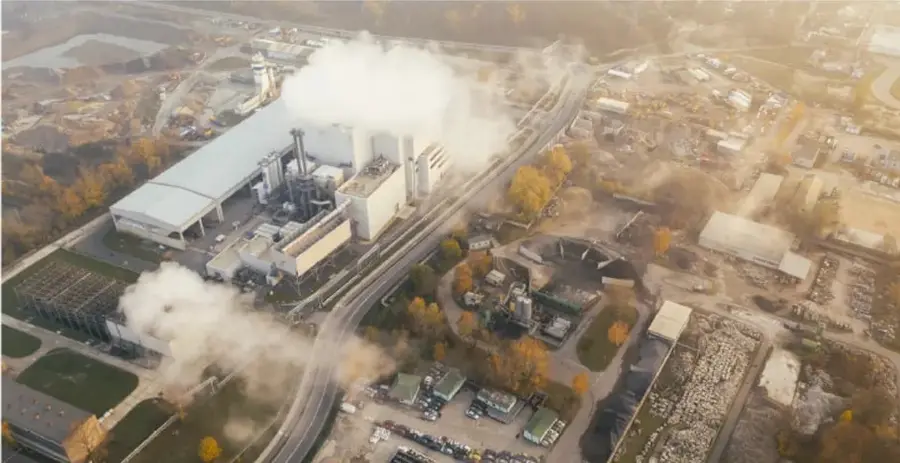 Vista aérea de una planta industrial con chimeneas que emiten humo blanco sobre un entorno urbano.