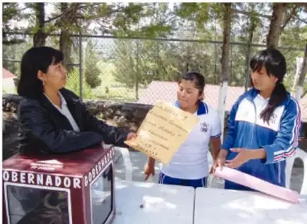 Fotografía de una funcionaria o autoridad entregando un documento a dos estudiantes frente a un módulo con la palabra 'GOBERNADOR'.