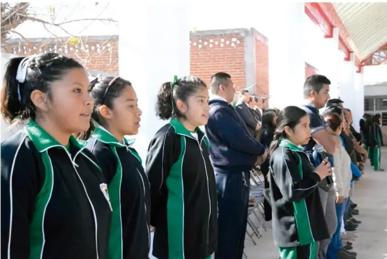Fotografía de estudiantes de telesecundaria con uniforme verde y negro, de pie en un patio escolar durante una actividad o ceremonia con asistentes adultos.