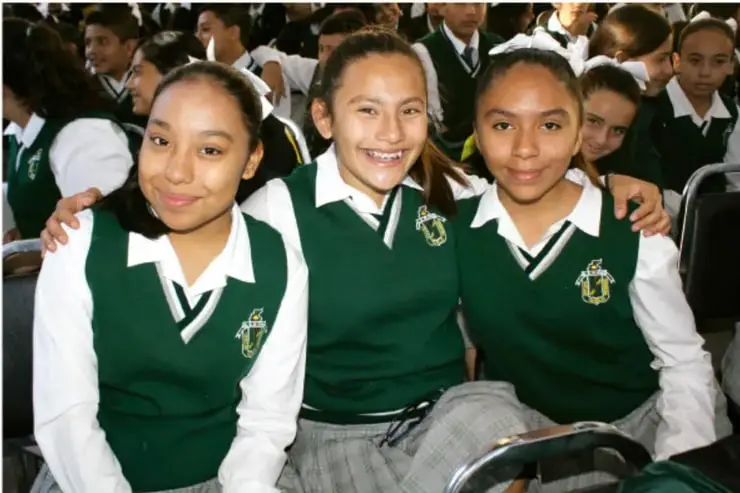 Fotografía de tres estudiantes con uniforme escolar verde y blanco, sonriendo y abrazándose, en un salón o auditorio con compañeros alrededor.