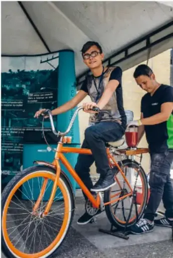 Dos jóvenes con una bicicleta naranja generando energía en un dispositivo tipo bicicleta generadora.