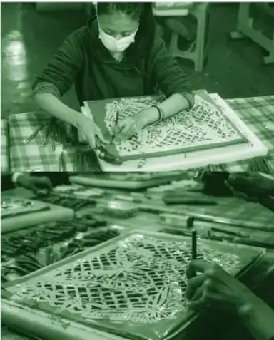 Fotografía a color (en tinta verde): una joven con cubrebocas talla papel picado sobre una mesa con cinceles, papel de china rosa y un patrón amarillo; otras manos sostienen la pieza abajo.