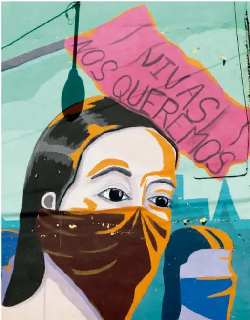 Mural urbano con el rostro de una mujer joven con cubrebocas naranja y negro; sobre ella una pancarta rosa con la frase 'VIVAS NOS QUEREMOS'. Al fondo, edificios y cables. Ilustra el arte como forma de expresión y denuncia social.