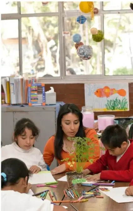 Fotografía de una maestra y tres estudiantes trabajando en una mesa con materiales escolares y una planta, en un aula con ventanal.
