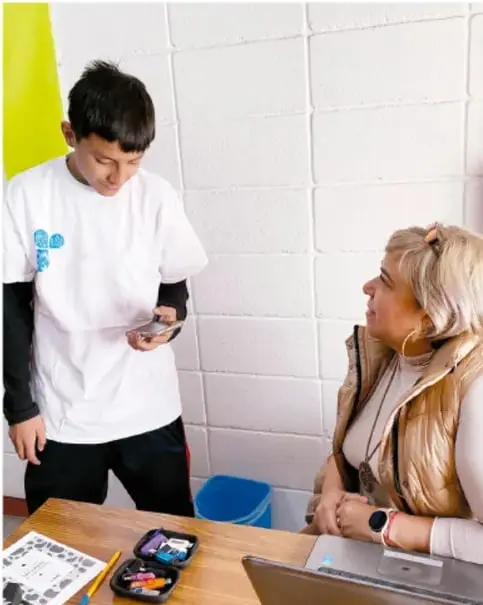 Foto de un estudiante de telesecundaria de pie mostrando un objeto o dispositivo a una docente sentada en un escritorio con una laptop y materiales escolares; ambos conversan en un aula.