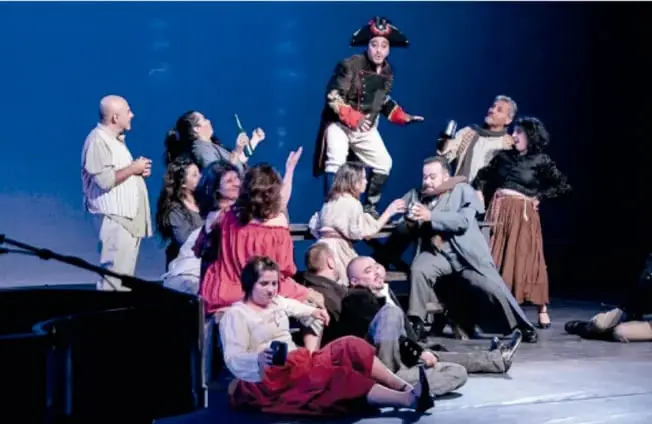Fotografía circular de una escena teatral: un actor mayor en silla de ruedas con traje, ante un telón con una gran rosa roja; al lado hay una figura encapuchada.