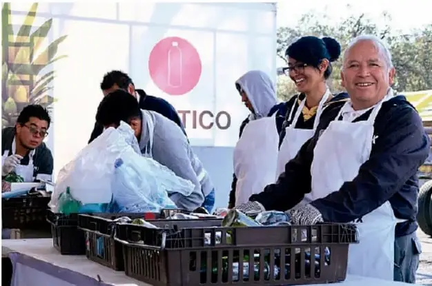 Fotografía de un trueque comunitario: varias personas con delantales blancos y guantes manipulan cajas y bolsas con objetos reciclables frente a un cartel con el símbolo de reciclaje y la palabra 'TICO'.