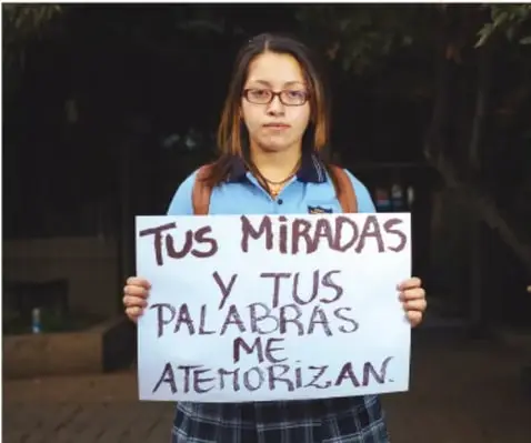 Fotografía de una joven con lentes y uniforme escolar que sostiene un cartel con la frase: "Tus miradas y tus palabras me atemorizan". Alude al acoso y a la violencia verbal.