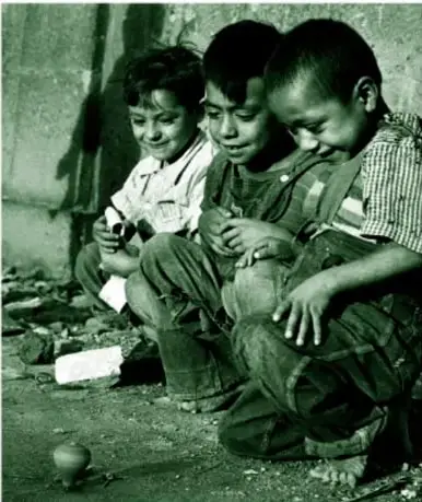 Fotografía 'Niños juegan con trompo' de Nacho López: tres niños en cuclillas, sonrientes, observan un trompo de madera girando sobre la tierra; en blanco y negro.