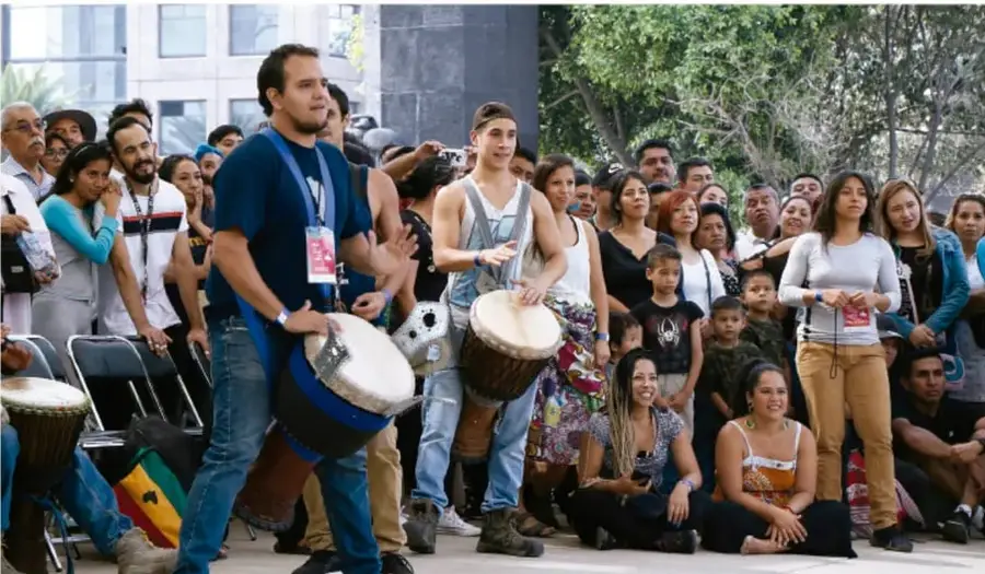 Grupo diverso de personas al aire libre participando en una actividad comunitaria con tambores; dos hombres jóvenes en el centro tocan instrumentos mientras demás personas observan y conviven.