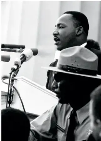 Fotografía en blanco y negro de Martin Luther King hablando frente a micrófonos, con una persona de sombrero en primer plano.