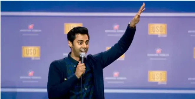 Fotografía de un comediante en el escenario del programa estadounidense "The USO Show", con micrófono y un brazo levantado, ante un fondo azul con el logo USO; se ilustra el humor en vivo.