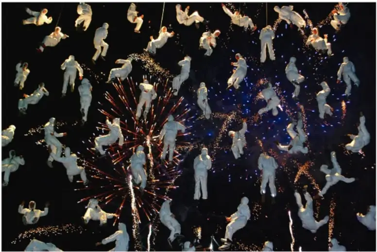 Fotografía nocturna de la compañía La Fura dels Baus: numerosos artistas vestidos de blanco suspendidos en el aire con arneses, rodeados de chispas y fuegos artificiales sobre fondo oscuro.